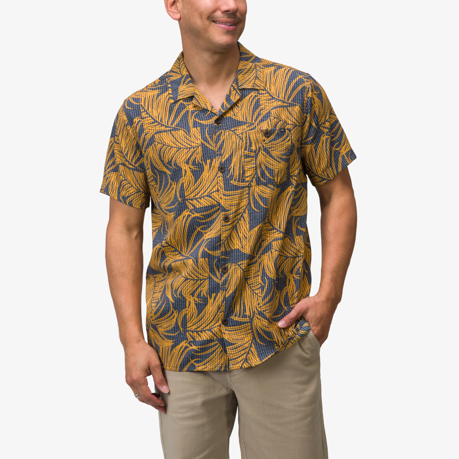 Man wearing a short-sleeve Kantis Shirt with blue and yellow leaf print, front view, hands in pockets, beach-inspired comfort apparel.