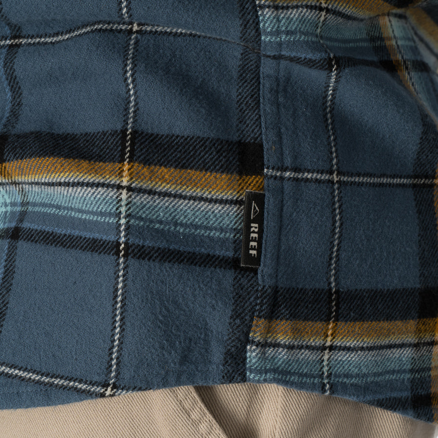 Close-up of the middle section of the blue and yellow plaid Dyson Flannel Shirt with a REEF tag visible on the side seam.