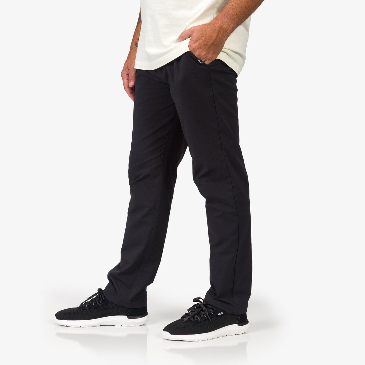 Men wearing black Carrick Stretch Pant facing left, paired with black REEF sneakers featuring anatomical arch support.