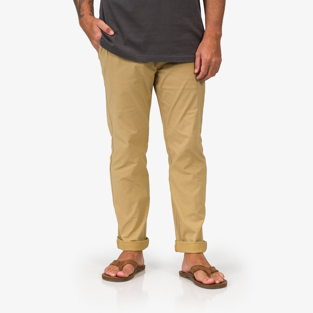 Front view of men's Carrick Stretch Pant in tan, paired with surf-inspired, comfortable brown sandals facing forward.