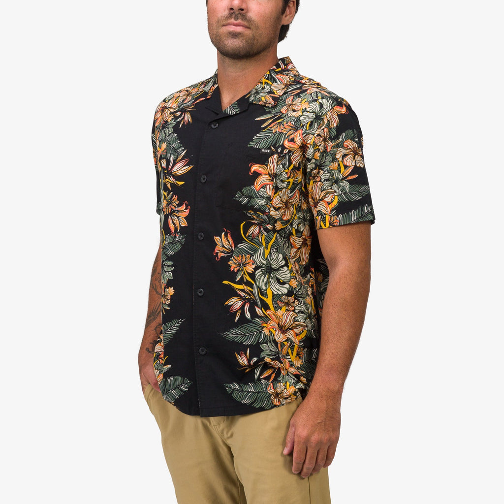 Man facing forward wearing Drift Ss Camp Shirt with orange and green floral pattern, hands in pockets, beige pants visible.