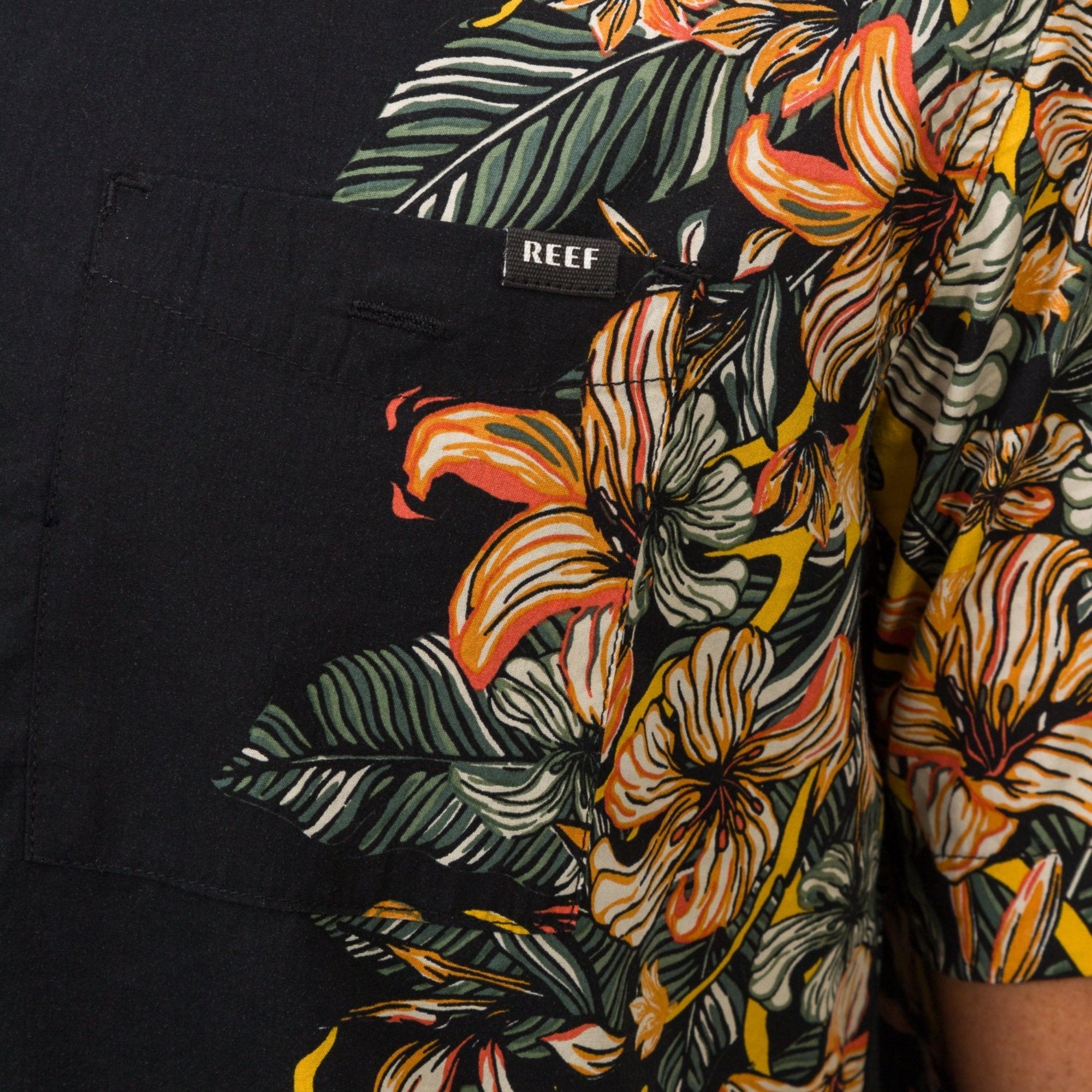 Close-up of Drift Ss Camp Shirt pocket and sleeve showing tropical floral print and Reef label on black fabric.