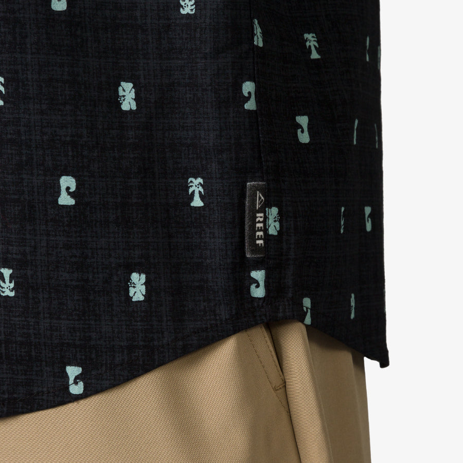 Close-up of the side hem on the black Chaplin Classic Shirt with light blue tiki pattern and REEF tag, worn with khaki shorts.