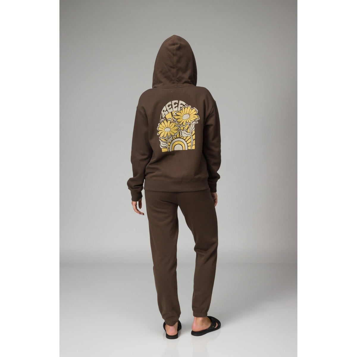 Person wearing brown Misty Fleece Full Zip hoodie back view with floral and rainbow design, paired with comfortable black sandals facing back