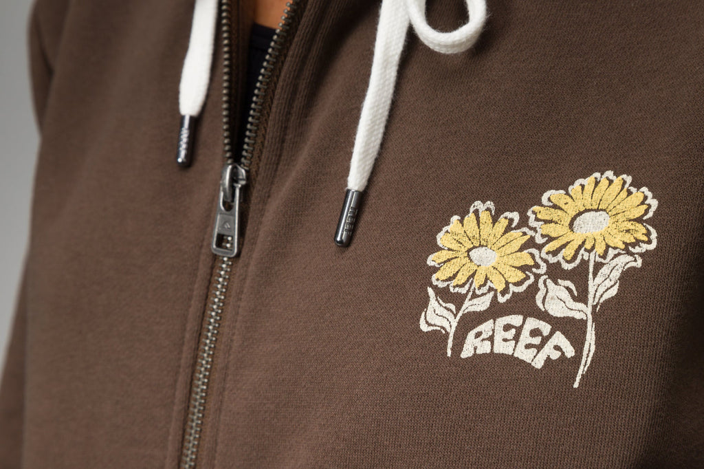 Close-up of chest area on Misty Fleece Full Zip showing embroidered yellow flowers and REEF logo on brown fabric with white drawstrings.