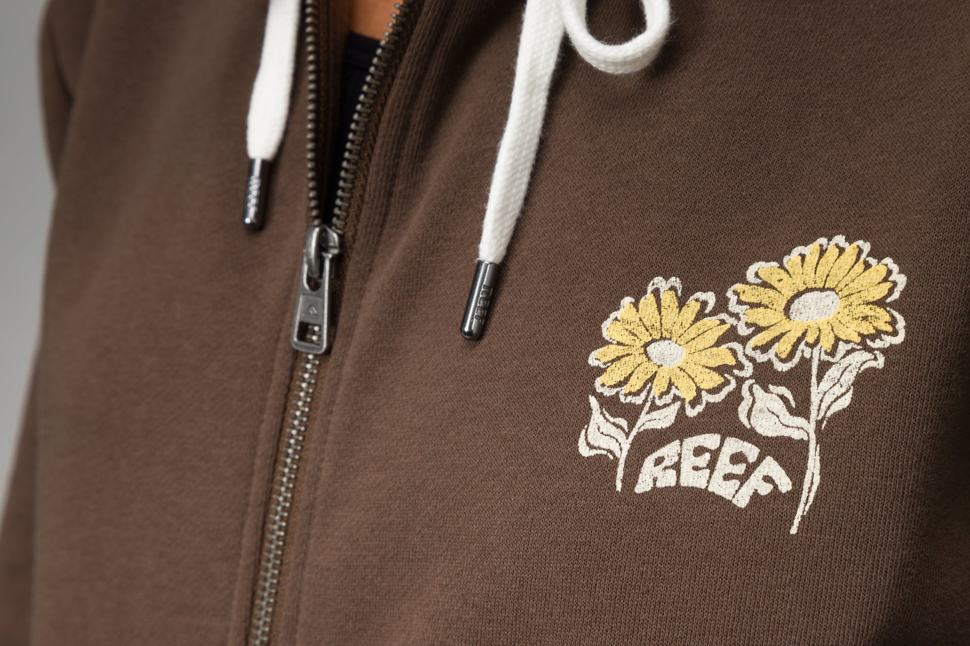 Close-up of chest area on Misty Fleece Full Zip showing embroidered yellow flowers and REEF logo on brown fabric with white drawstrings.
