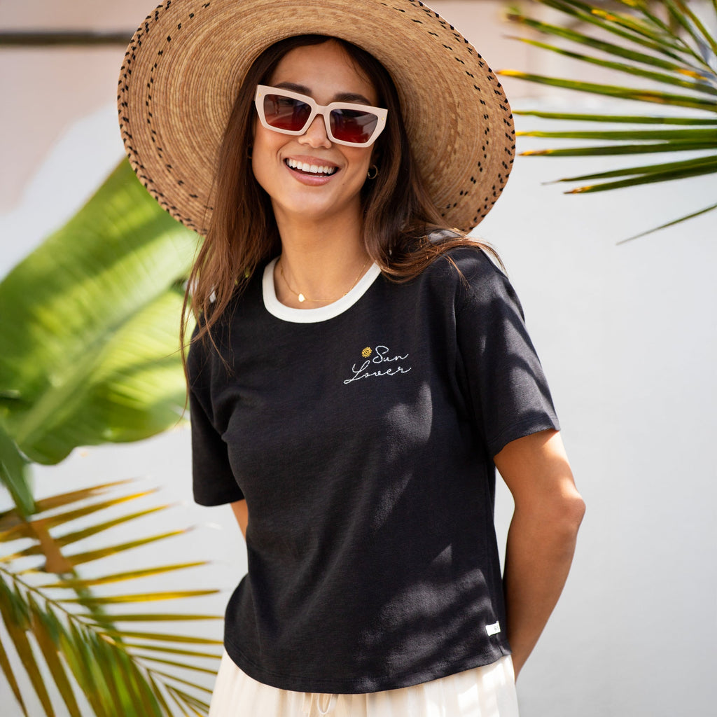 Woman wearing a black Sun Lover Ringer Tee with white collar and embroidered logo, smiling outdoors with a large sun hat.