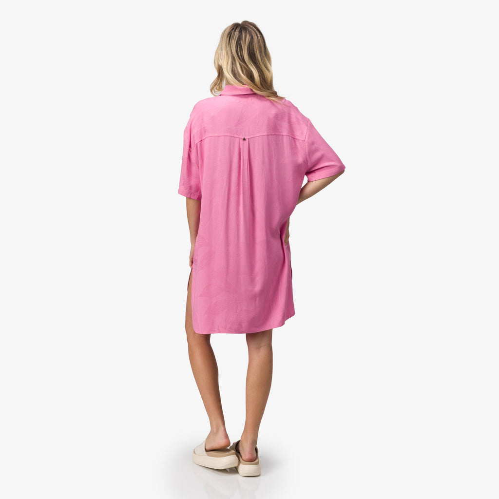 Woman wearing a pink Ollie Shirt Dress shown from the back, paired with beige sandals facing left, beach-inspired comfort style.