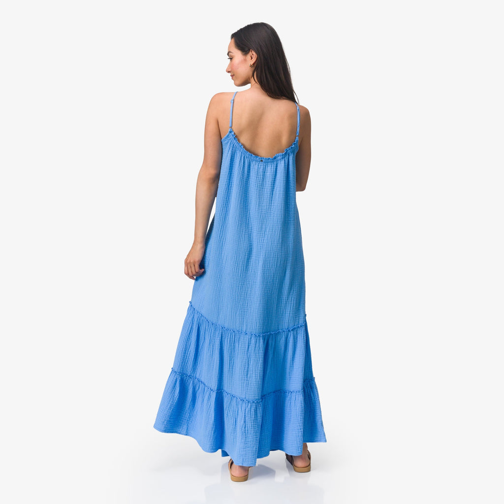Woman wearing a blue Lainey Maxi Dress shown from back with thin straps, flowing tiers, and casual sandals facing right.