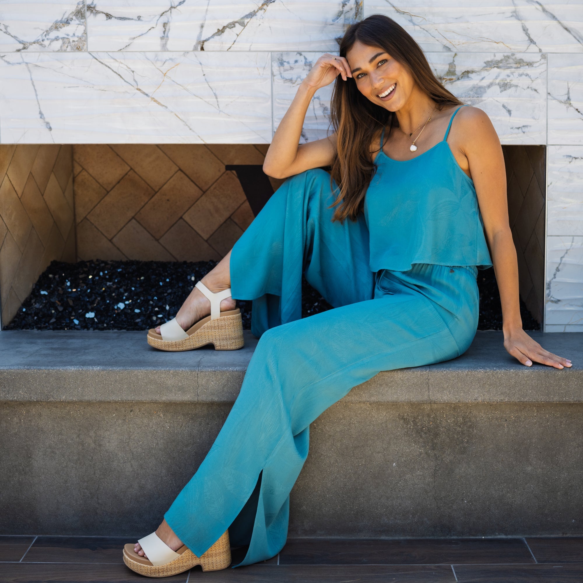 Woman wearing a turquoise Ollie Tank sitting outdoors, smiling, showing front and side of platform sandals facing right