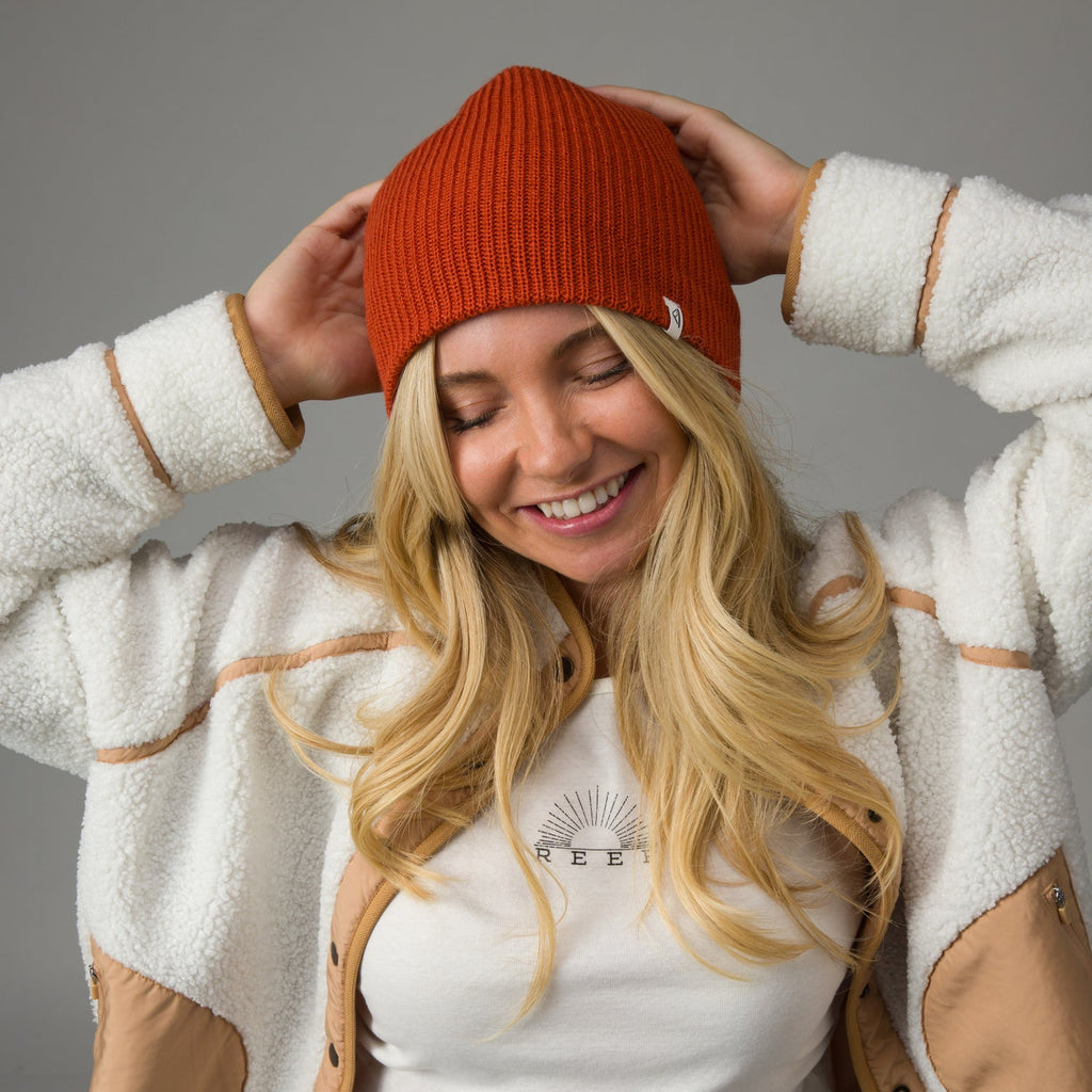 Smiling woman wearing a chili red Alyx Beanie, front view, styled with cozy fleece jacket and casual beachwear.