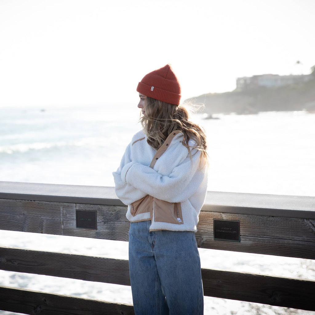 Woman wearing a chili colored Alyx Beanie facing left near ocean, styled with casual beachwear and denim pants
