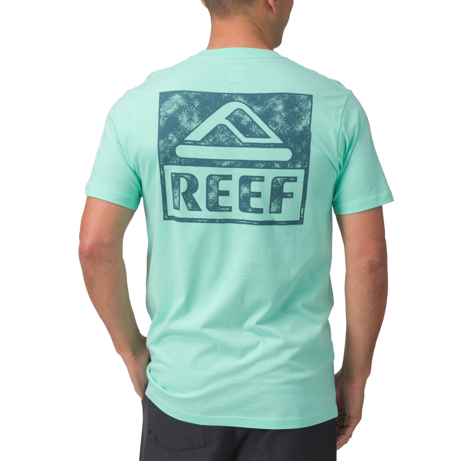 Man wearing light blue Wellie Too Tee facing away showing back logo, casual surf-inspired apparel for the beach.