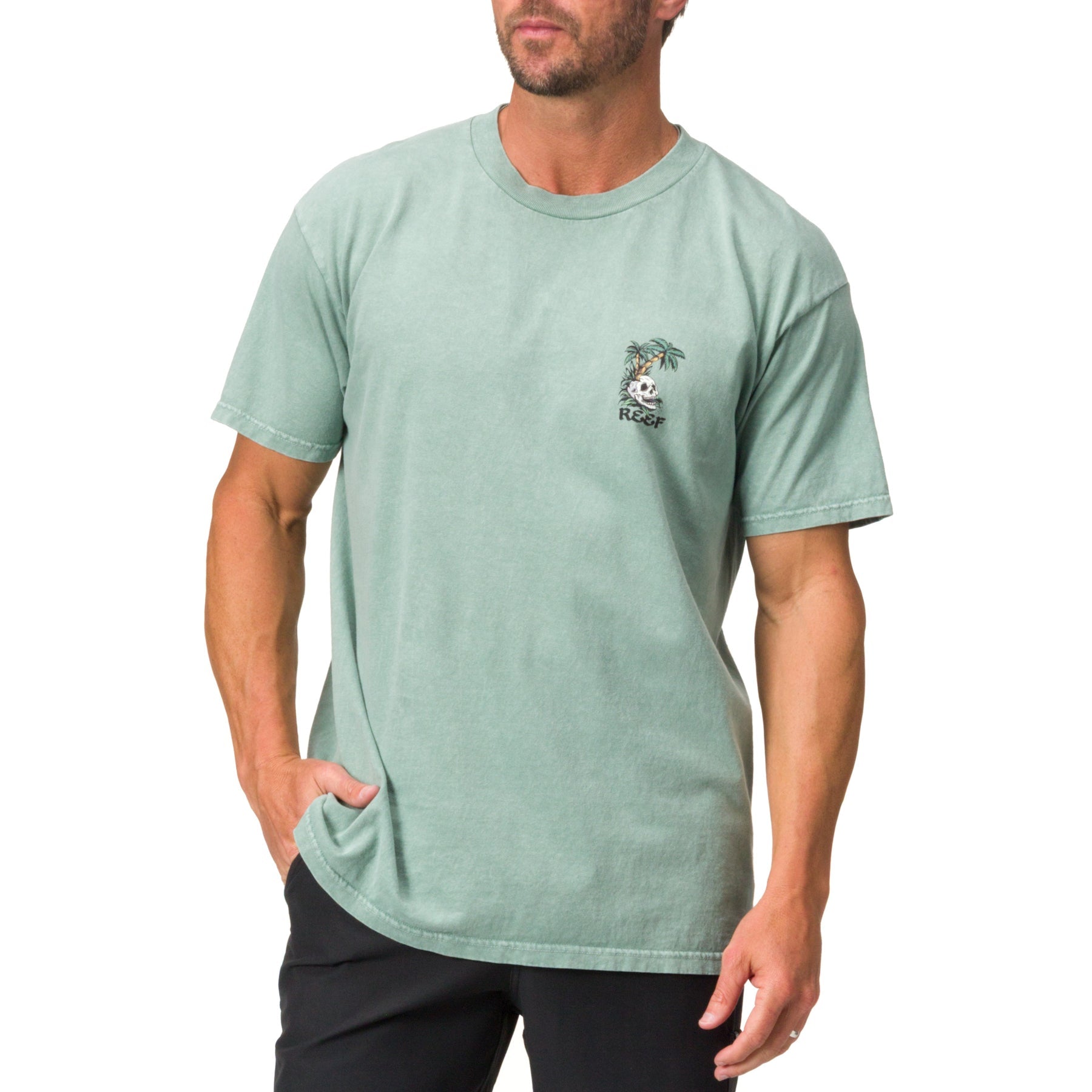 Front view of a man wearing a green Akin Tee with a small palm tree graphic, surf-inspired casual apparel for men.