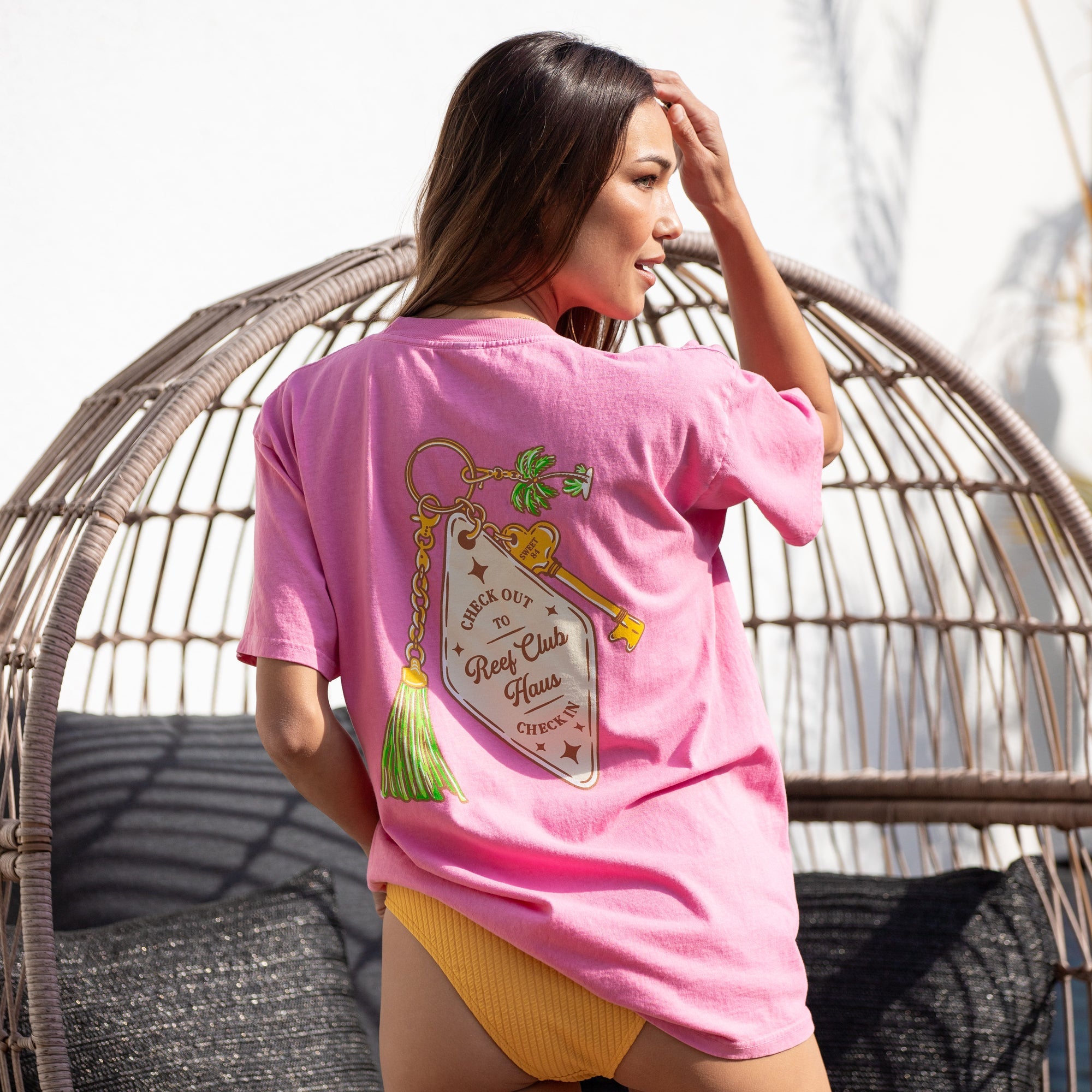 Woman wearing pink Haus Boyfriend Crew shirt facing away, highlighting back graphic, seated outdoors in casual beachwear.