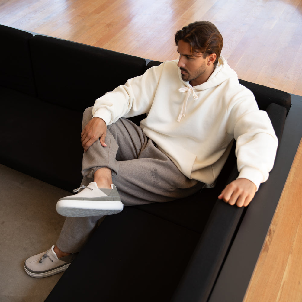 Man sitting on black couch wearing white hoodie and gray pants with Rambler shoes facing left, highlighting comfort and style.