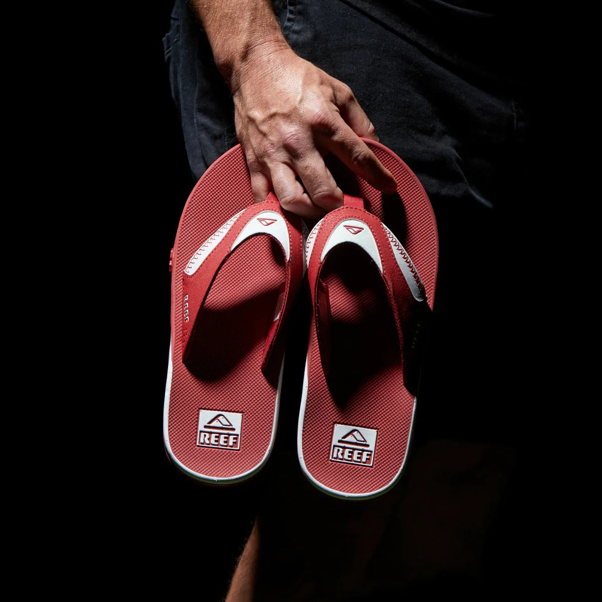 Hand holding a pair of red and white Fanning Pregame flip flops facing sole up showing cushioned arch support and REEF logo