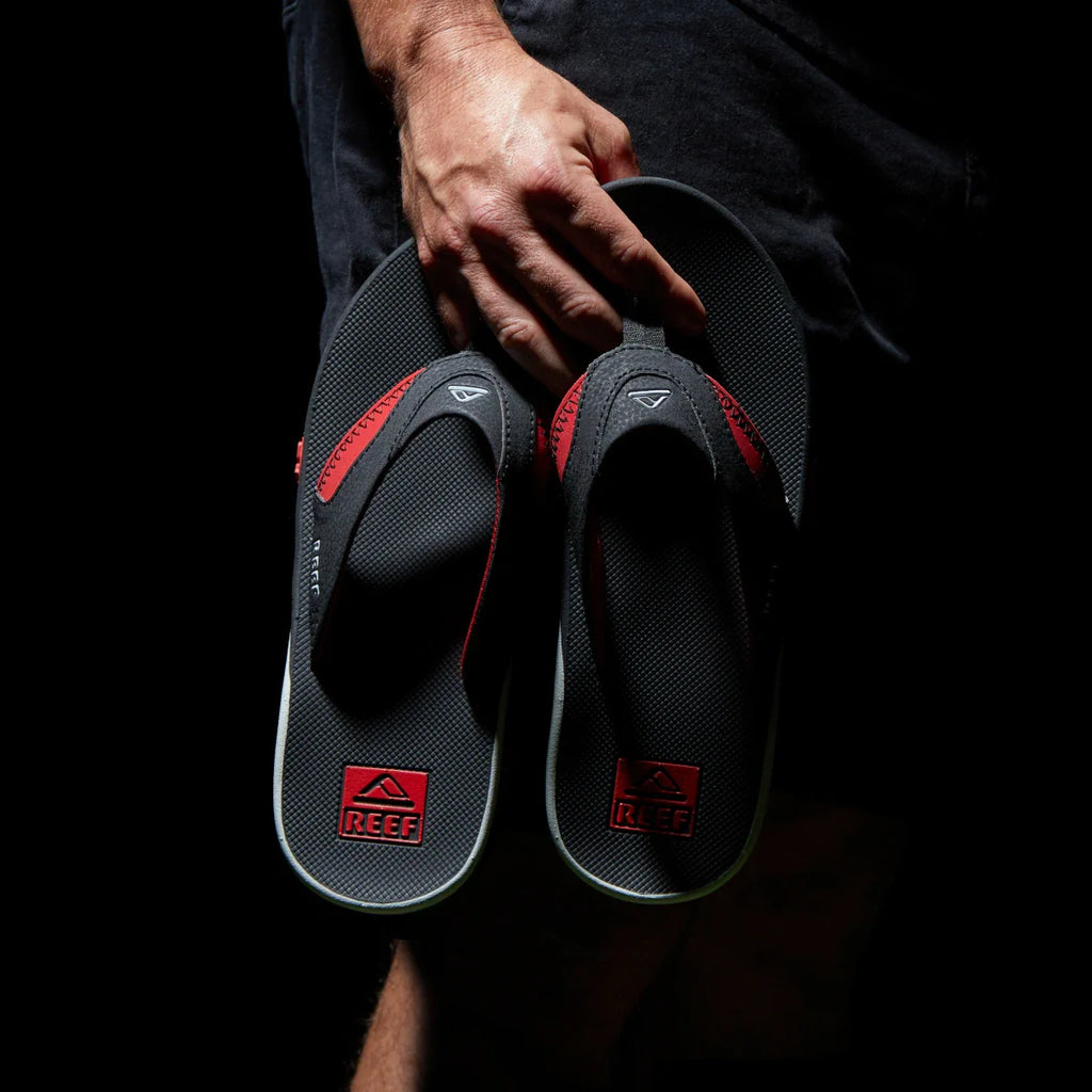 Hand holding a pair of Fanning Pregame men's black and red flip flops, showing front straps and soles facing forward.