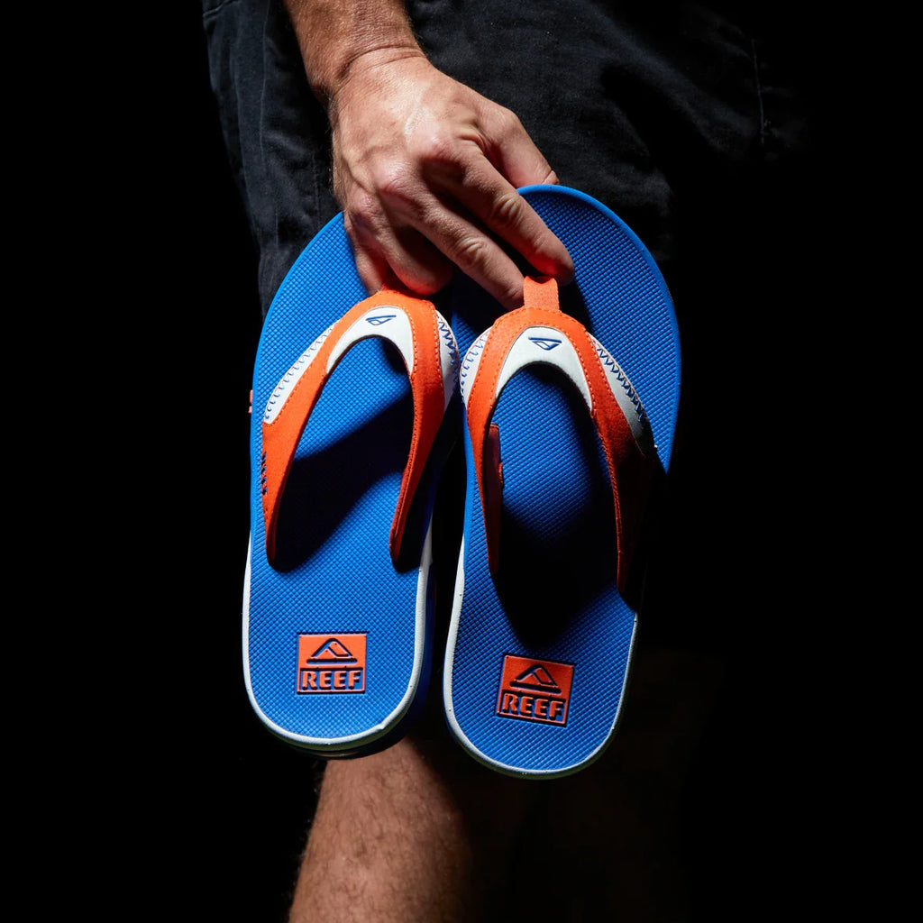 Hand holding a pair of Fanning Pregame flip flops facing up, with blue soles and orange straps, beach-inspired comfort.