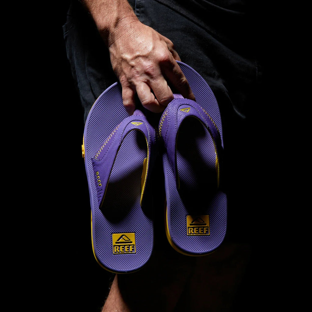 Hand holding purple and gold Fanning Pregame flip flops facing up, showcasing soft-rebound cushioning and arch support.