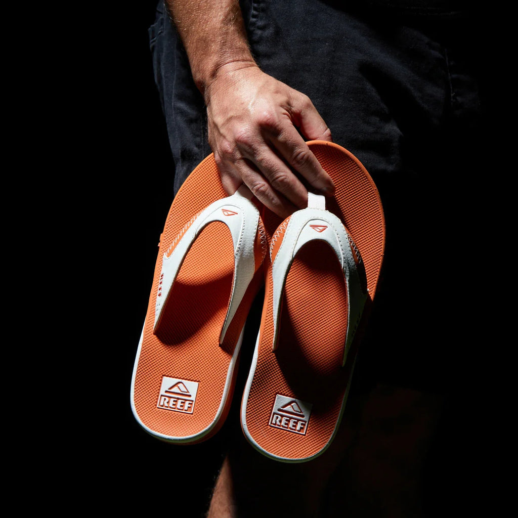Hand holding a pair of men's Fanning Pregame flip flops facing sole up, showing orange footbed and white straps with arch support.