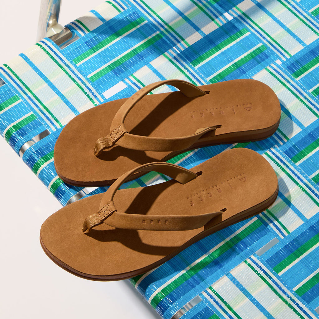 Pair of brown Ojai Classic Slim sandals facing left on a striped blue and green beach chair, showing top and side view.