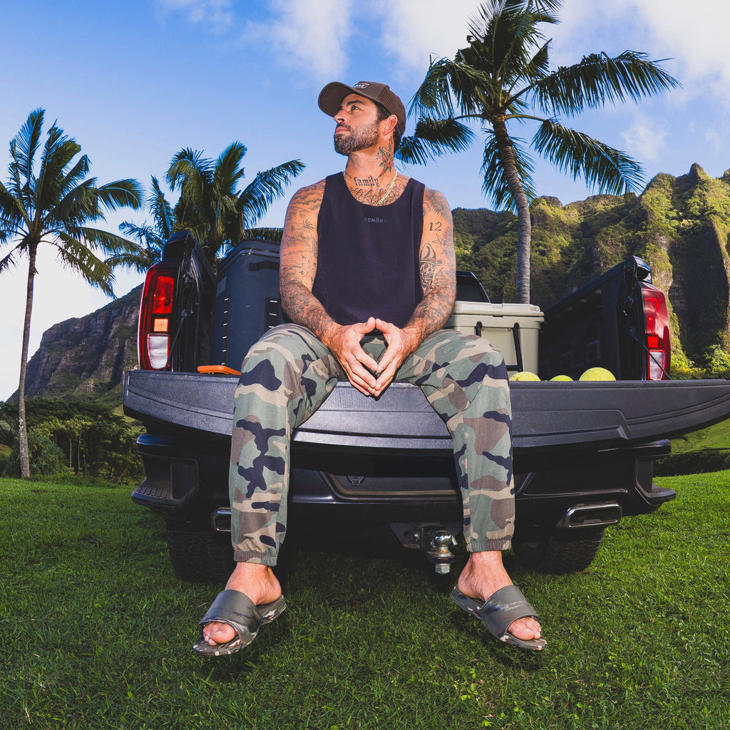 Man wearing camo Fanning Slide H2O facing forward, seated on truck tailgate with tropical palm trees in background