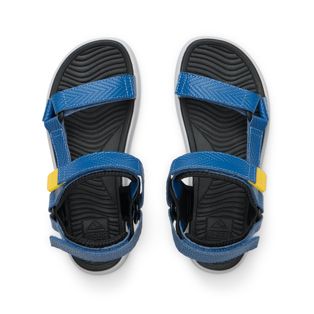 Kids Shoremazing sandals facing upward, blue straps with yellow details, black textured footbeds, beach-inspired comfort design.