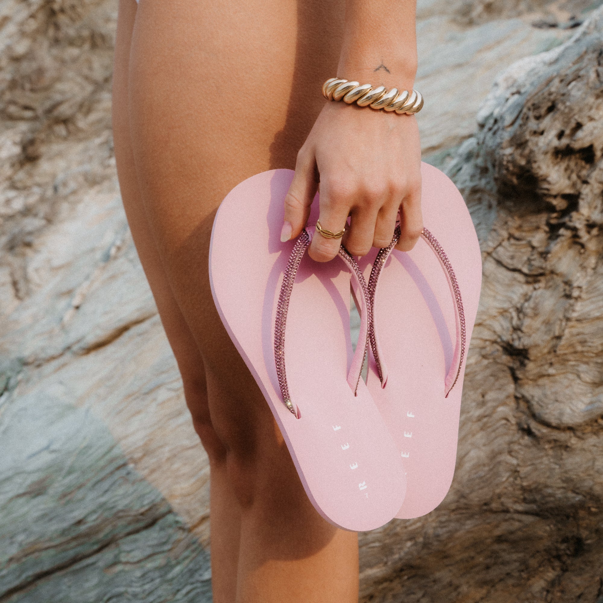 Woman holding pink Bliss Bling Fling sandals by the straps, showcasing comfort and beach-inspired design.