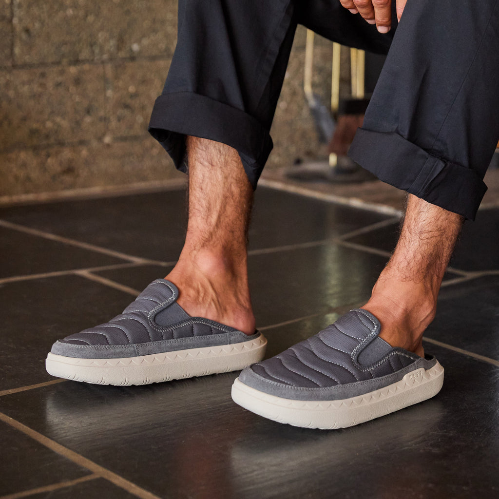 Rambler Kicker slippers facing right worn indoors, featuring cushioned memory foam footbed and lightweight EVA outsole in charcoal gray