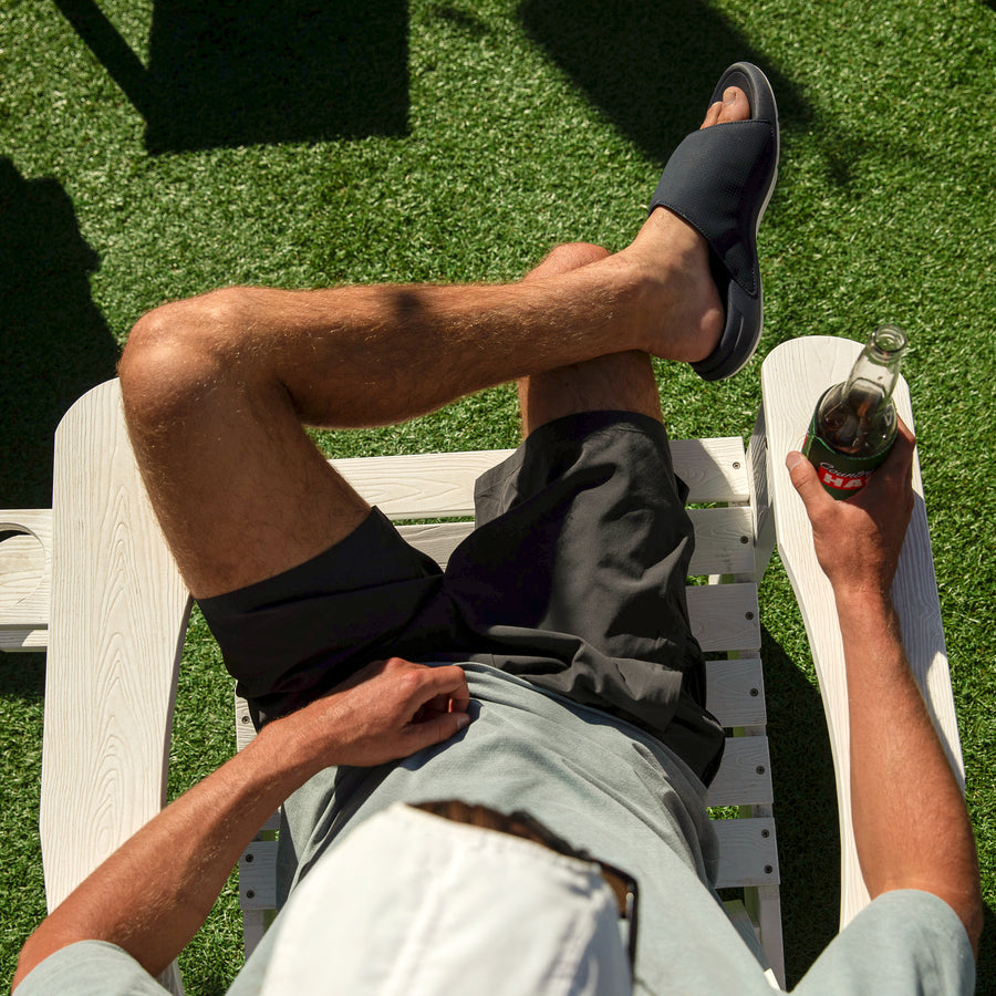 Man relaxing on white chair wearing navy and white Cruiser Slide on right foot facing right showing side view with drink in hand