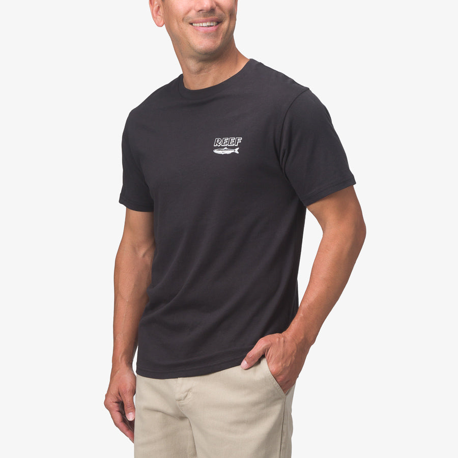 Man wearing black Cannery Tee with small white REEF logo on chest, posing with hand in pocket, beach-inspired comfort apparel.