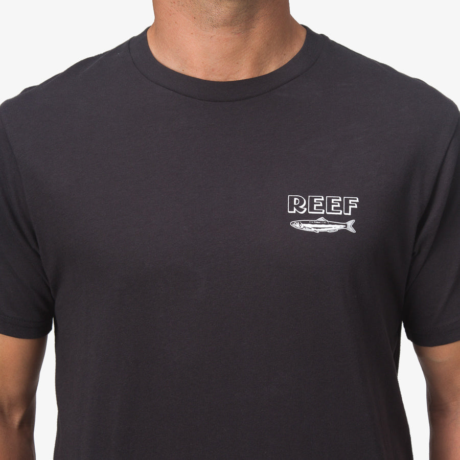 Close-up of a man wearing a black Cannery Tee with a small REEF logo and fish graphic on the chest, surf-inspired comfort apparel.