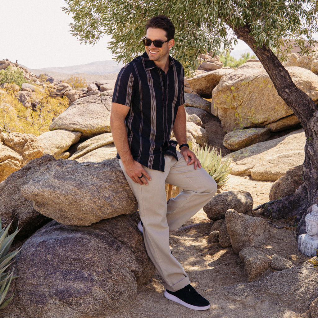 Man wearing Carrick Stretch Pant in beige sitting on rocks outdoors, black footwear facing left, casual beach-inspired style