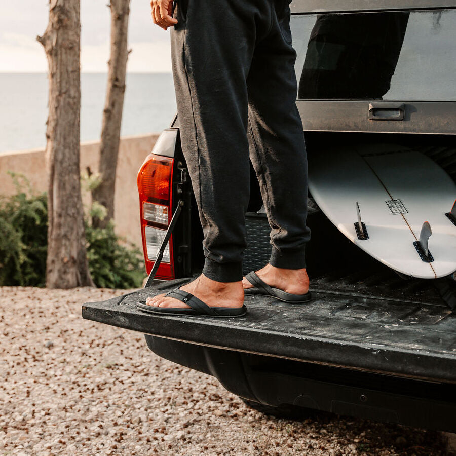 Cushion Phantom 2.0 sandals facing right on adult male feet, shown from side near vehicle tailgate and surfboard.