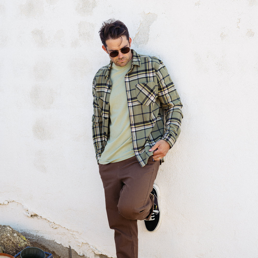 Man wearing Dyson Flannel Shirt in olive plaid, facing left, styled for beach and active lifestyles with sunglasses and casual pants.