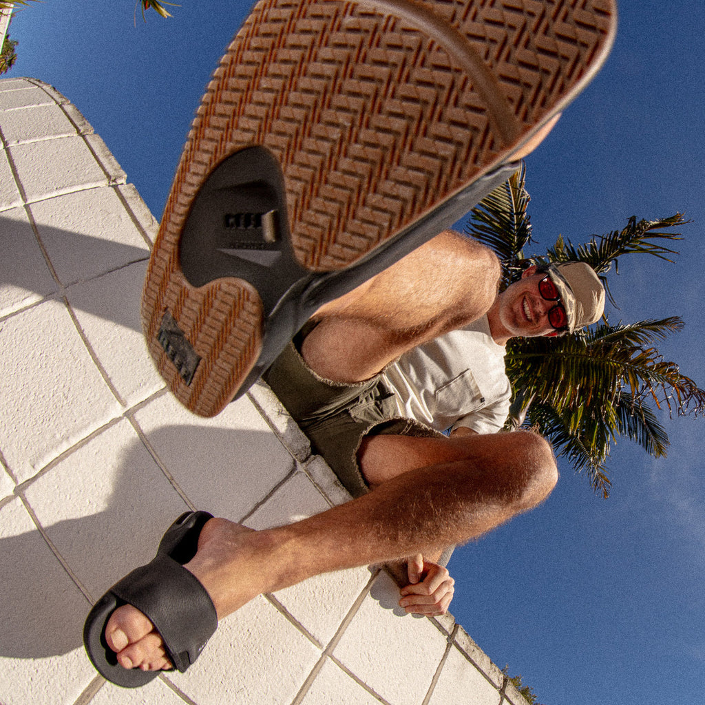 Fanning Slide sandal in black facing forward, close-up on sole with man wearing and sitting outdoors by palm trees.