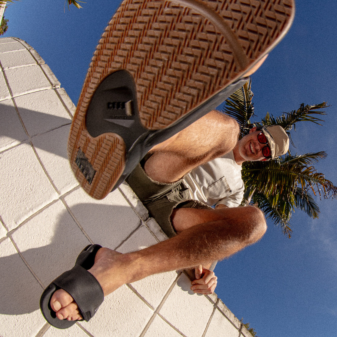 Fanning Slide sandal in black facing forward, close-up on sole with man wearing and sitting outdoors by palm trees.
