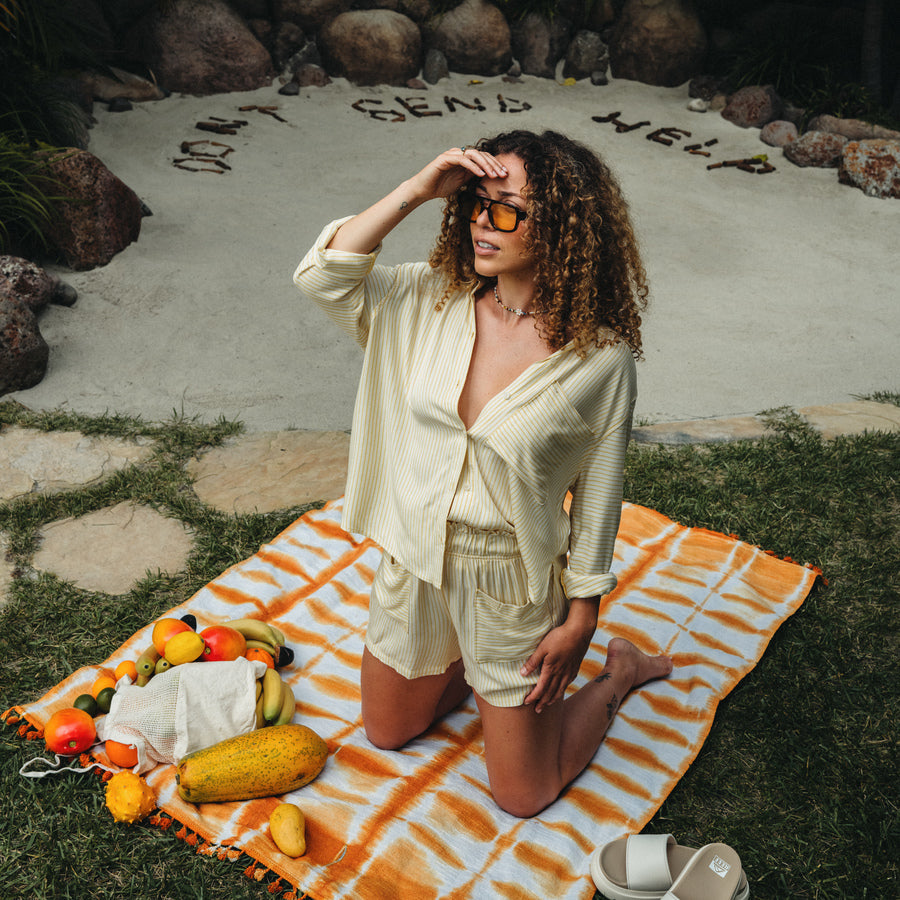 Woman wearing Ellie Button Up cream striped shirt kneeling on beach towel with fruits nearby, shaded by her hand