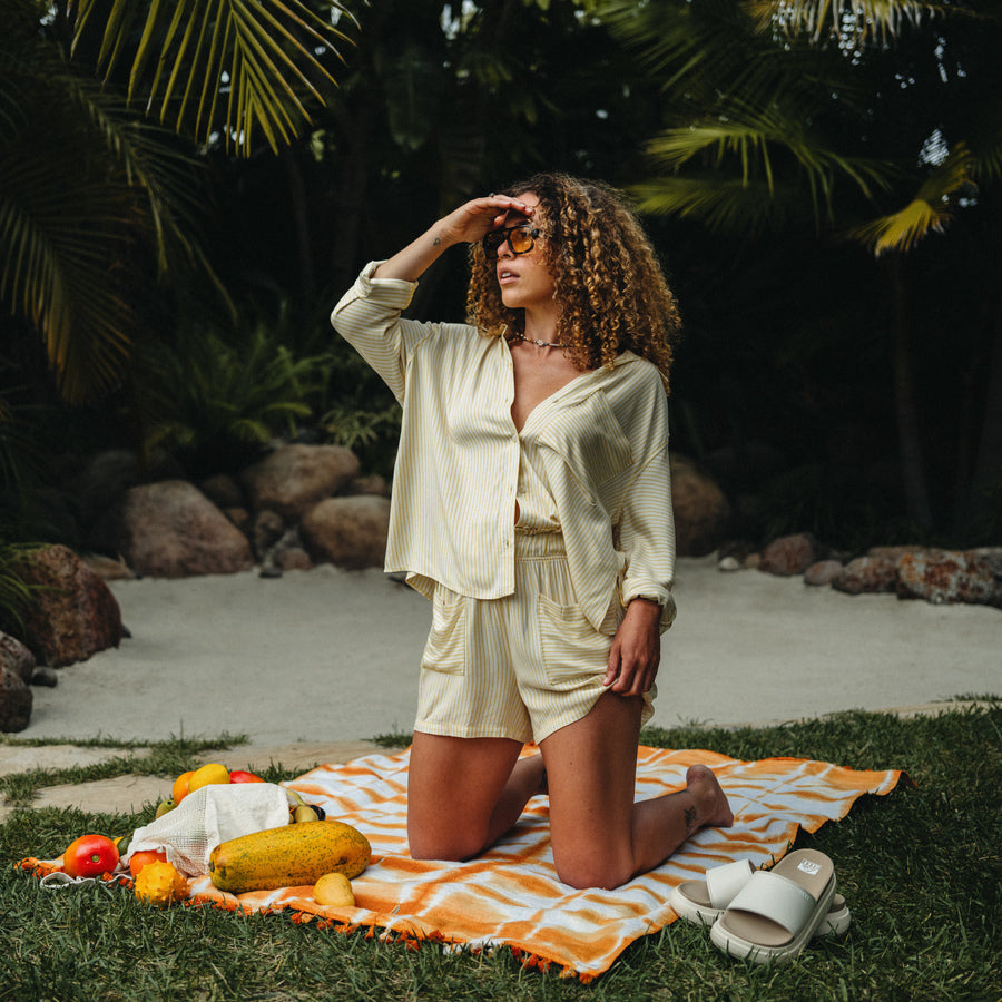 Woman wearing Ellie Short kneeling on a beach towel with side-facing white REEF sandals beside her outdoors
