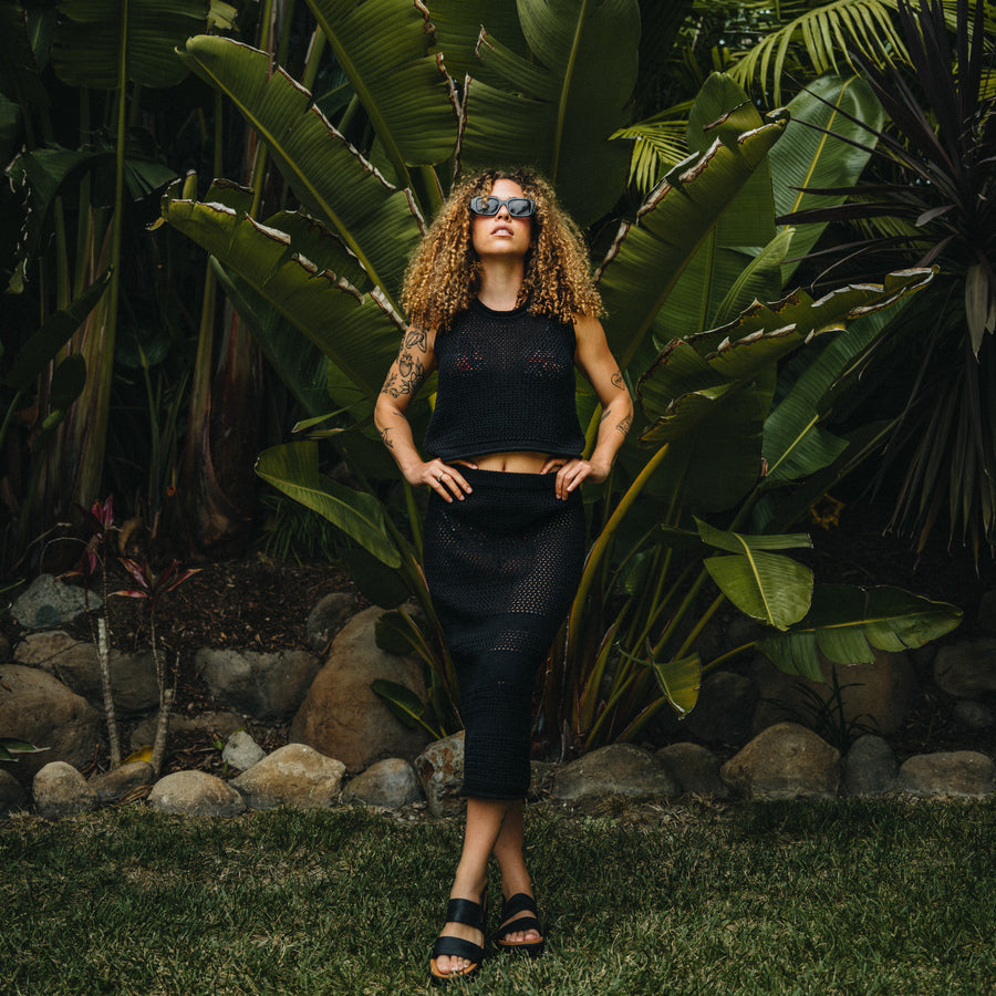 Woman wearing Mika Sweater Tank in black, standing outdoors with hands on hips, wearing black sandals facing forward.