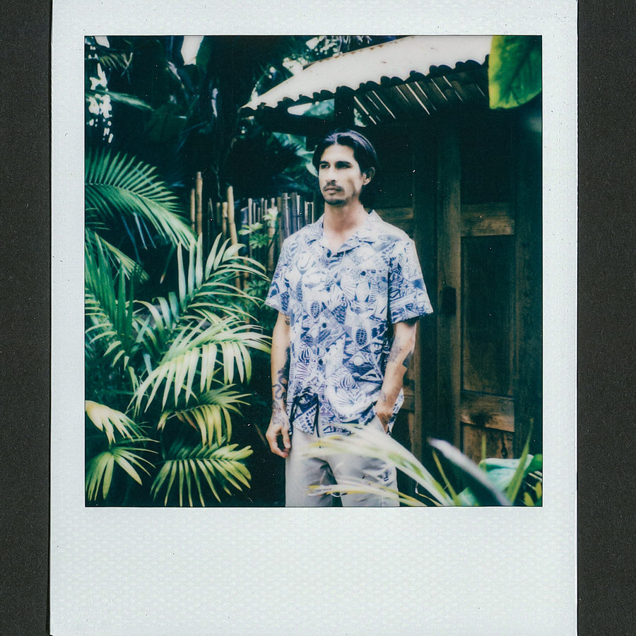 Man wearing a blue patterned Beckham Camp Shirt standing outdoors by tropical plants and wooden structure in relaxed pose