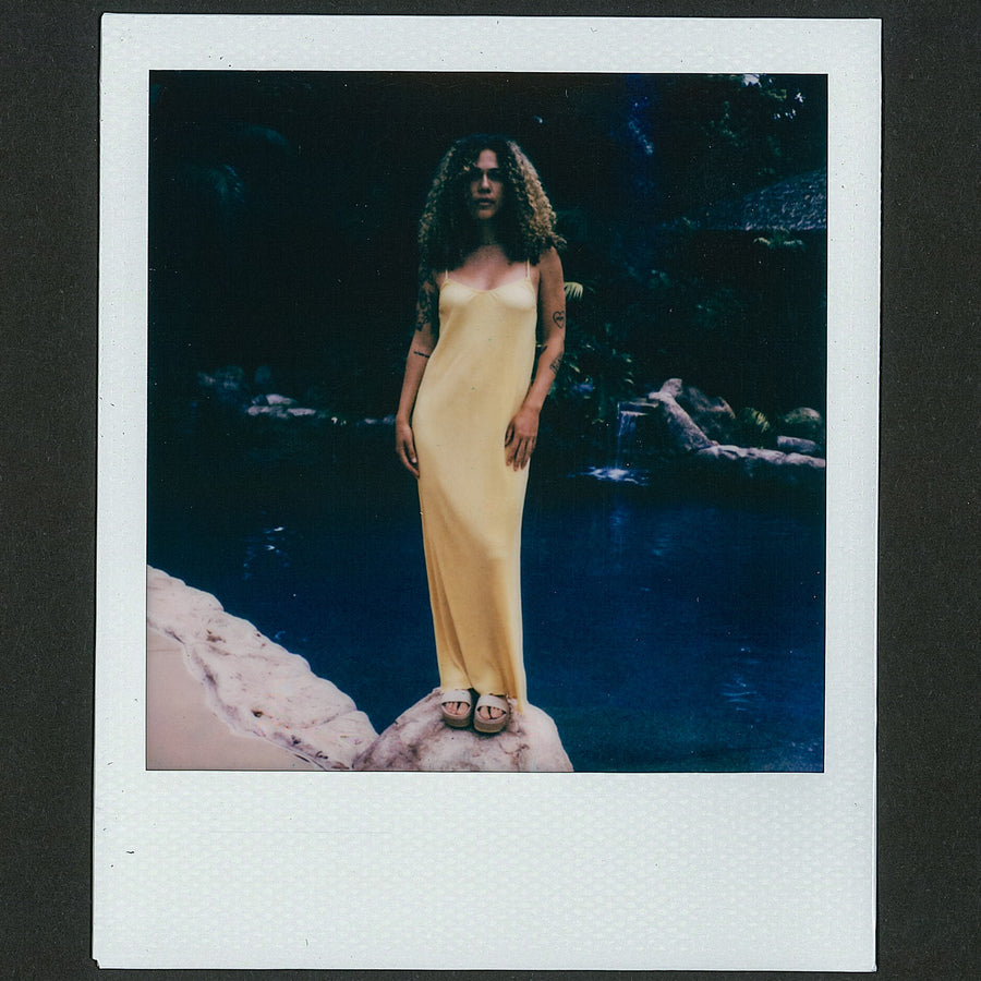 Woman wearing a yellow Plisse Dress standing near a rocky pool, showcasing surf-inspired beach apparel and comfort sandals facing forward.