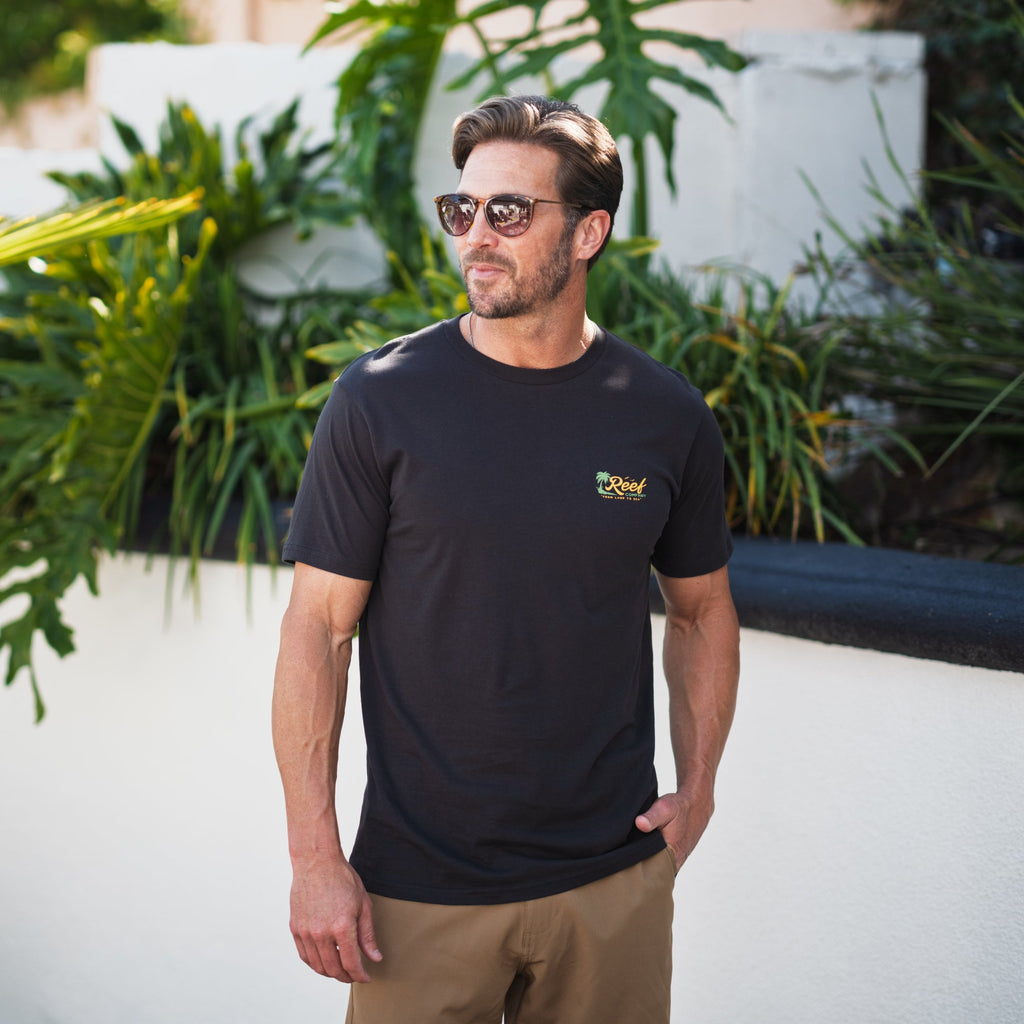 Man wearing a black Isac Tee with sunglasses, standing outdoors near greenery, showcasing relaxed beach-inspired comfort.