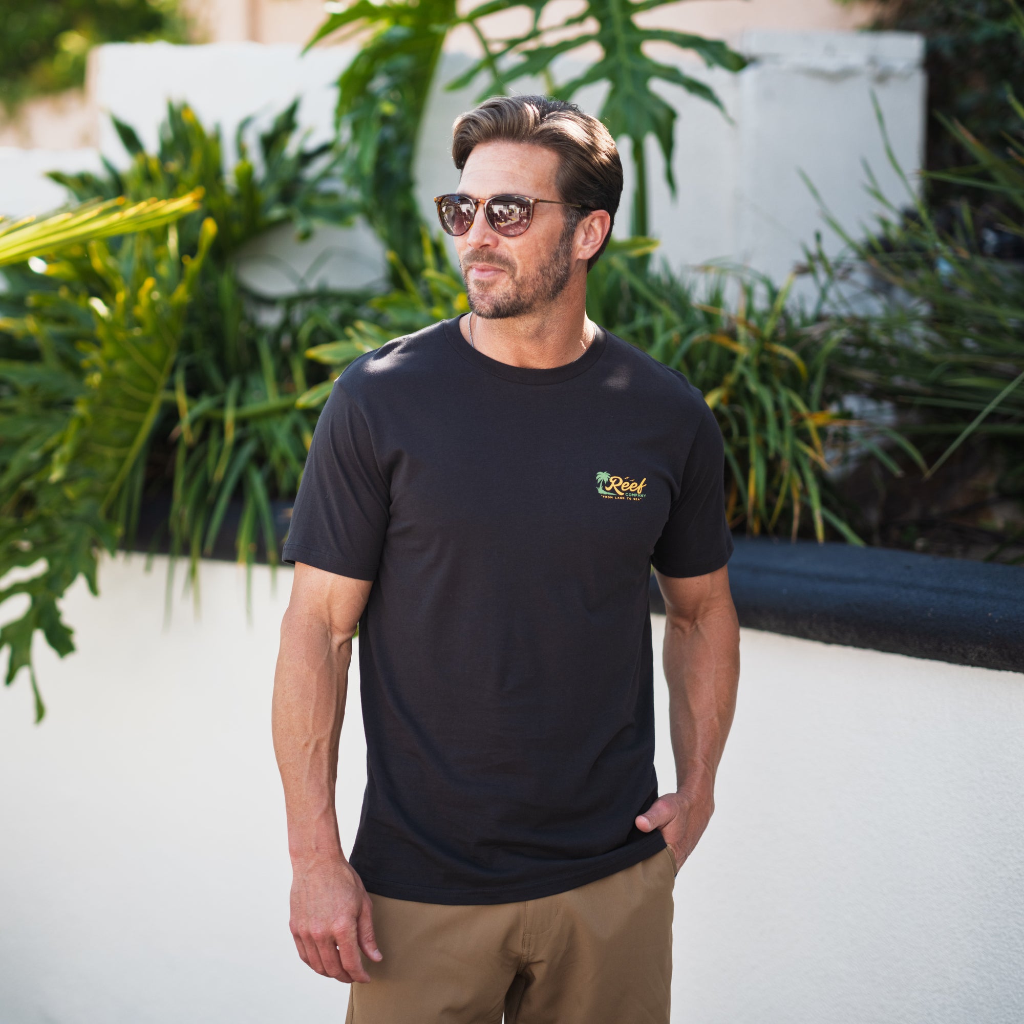 Man wearing a black Isac Tee with sunglasses, standing outdoors near greenery, showcasing relaxed beach-inspired comfort.