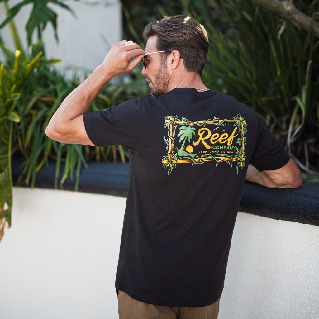 Man wearing black Isac Tee showing back graphic design outdoors, beach-inspired comfort apparel for men.