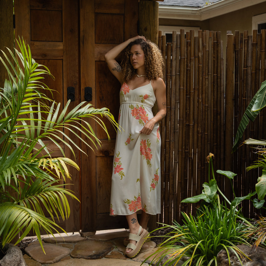 Woman wearing Belladona Dress with floral print, standing outdoors near wooden door and bamboo fence, sandals visible facing forward.