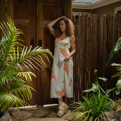 Woman wearing Belladona Dress with floral print, standing outdoors near wooden door and bamboo fence, sandals visible facing forward.