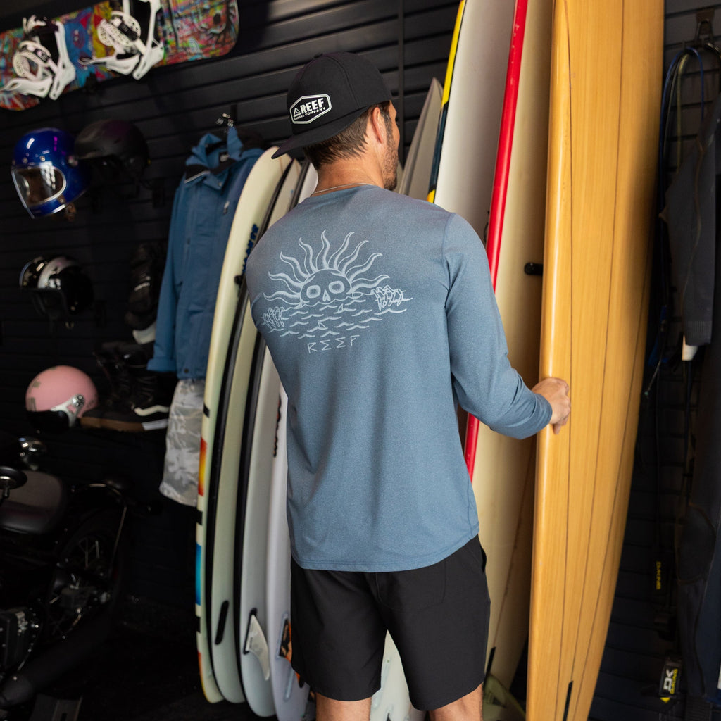 Man wearing a blue Set In Performance Shirt facing away, holding a surfboard in a surf shop with beach gear.