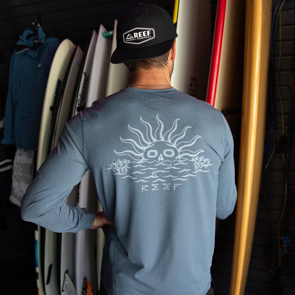 Man wearing a blue Set In Performance Shirt showing back design, standing by surfboards in a beach-inspired setting